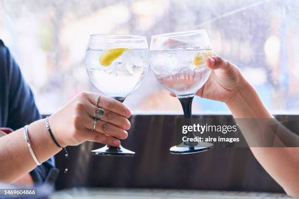 two people holding glasses with gin cocktail, lemon strip with ice, one of the hands has rings and bracelets in the background glass wall. - gin tónico - fotografias e filmes do acervo