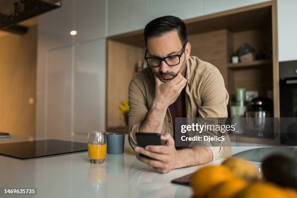 pensive young man using his cell phone on kitchen counter at home. - s thirtysomething stock pictures, royalty-free photos & images