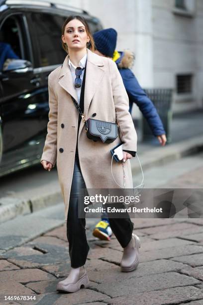 Guest wears a white shit, black sunglasses, a beige wool coat, a black shiny leather embossed eyes pattern crossbody bag from Chiara Ferragni, black...