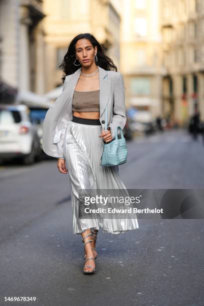 Ciinderella Balthazar wears silver earrings, a silver necklace, a pale gray blazer jacket, a brown V-neck cropped t-shirt, a pale blue shiny leather...