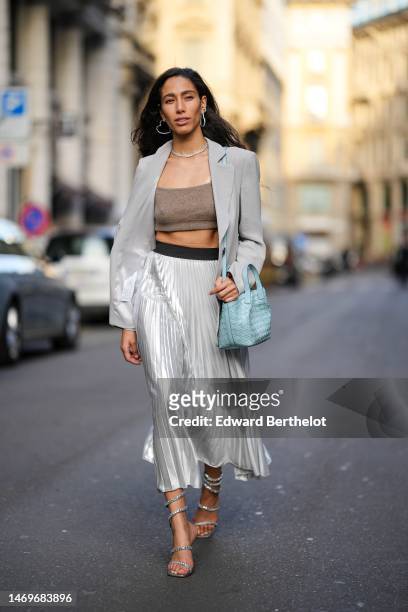 Ciinderella Balthazar wears silver earrings, a silver necklace, a pale gray blazer jacket, a brown V-neck cropped t-shirt, a pale blue shiny leather...