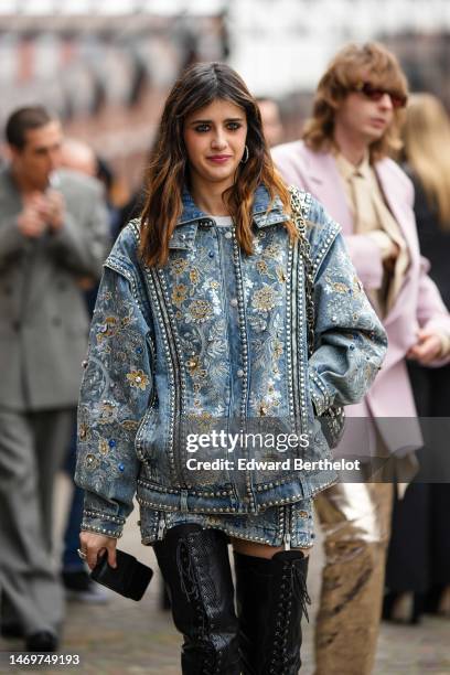 Benedetta Porcaroli wears silver earrings, a blue denim with embroidered gold and silver flower pattern oversized bomber jacket, a matching blue...