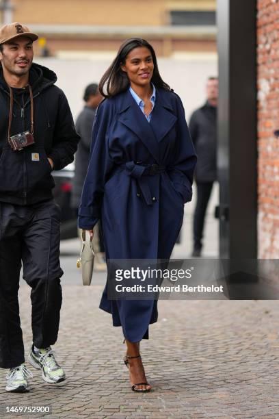 Tina Kunakey wears a pale blue shirt, a navy blue belted long trench coat, a white latte leather handbag from Gucci, black shiny leather strappy...