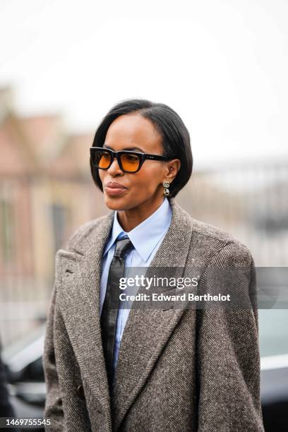 Sabrina Dhowre Elba wears black and orange sunglasses, silver pendant logo earrings from Gucci, a pale blue shirt, a black shiny leather tie, brown...