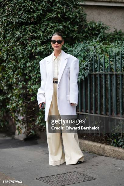 Sophia Roe wears black sunglasses, a white latte shirt, a white oversized blazer jacket, a black shiny leather belt, pale yellow large wide legs suit...