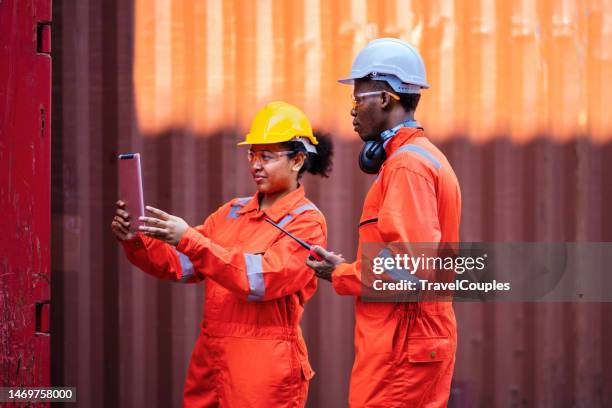 multiethnic female industrial engineer with tablet and black african american male supervisor in hard hats and safety vests stand in container terminal. colleagues talk about logistics operations. business logistic concept. import and export concept - team talk stockfoto's en -beelden