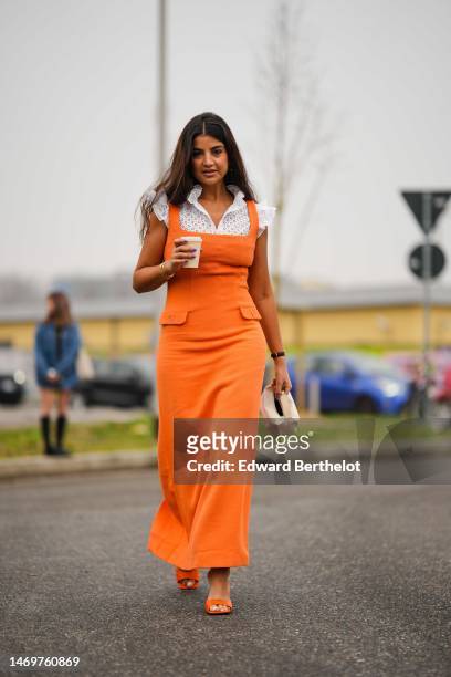 Ola Farahat wears gold earrings, a white cut-out flower pattern ruffled short sleeves blouse, an orange squared-neck / tank-top long tube dress, a...