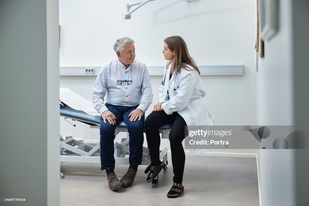 Mature patient talking to doctor while sitting on bed in clinic