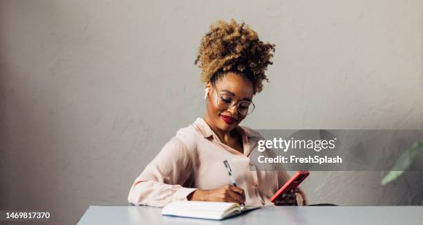 a happy beautiful businesswoman with blonde curly hair taking some notes while using her earbuds and mobile phone - curly red hair glasses stock pictures, royalty-free photos & images