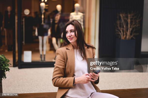 elegante donna bruna che distoglie lo sguardo mentre tiene il suo telefono cellulare in un centro commerciale all'aperto in cappotto marrone. - trench foto e immagini stock