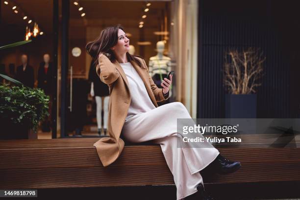 donna alla moda in un cappotto di lana marrone alla moda seduta all'aperto mentre si prende una pausa da un lavoro in ufficio. - trench foto e immagini stock