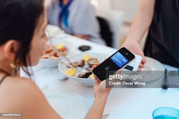 young asian woman using contactless payment with smartphone to pay for a meal at restaurant - apple pay stock pictures, royalty-free photos & images