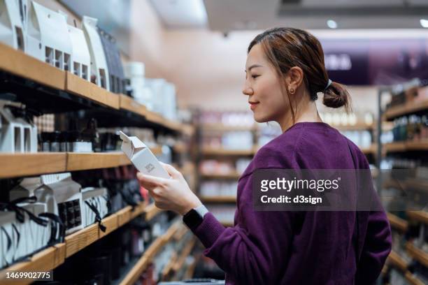 young asian woman shopping for natural beauty products in organic health and beauty store. looking at the ingredients of the skincare package. personal skincare products. health and wellness concept. sustainable lifestyle - femme make up mauve photos et images de collection