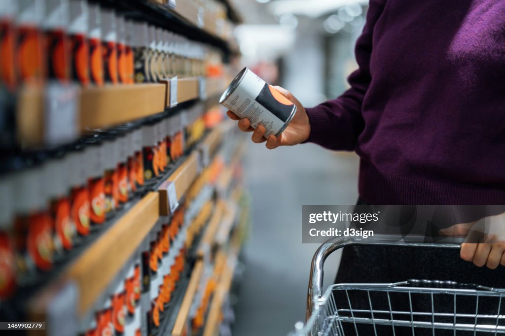 Cropped shot of young woman pushing a shopping cart, grocery shopping in supermarket. She is holding a tin can and reading the nutritional label. Routine grocery shopping. Food shopping. Making healthier food choices