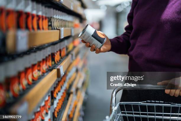 cropped shot of young woman pushing a shopping cart, grocery shopping in supermarket. she is holding a tin can and reading the nutritional label. routine grocery shopping. food shopping. making healthier food choices - producto enlatado fotografías e imágenes de stock