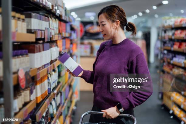 young asian woman pushing a shopping cart, grocery shopping in supermarket, picking up a box of coffee beans from the shelf. routine grocery shopping. food shopping. making healthier food choices - coffee package stock-fotos und bilder