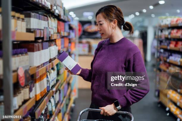 young asian woman pushing a shopping cart, grocery shopping in supermarket, picking up a box of coffee beans from the shelf. routine grocery shopping. food shopping. making healthier food choices - label coffee bildbanksfoton och bilder