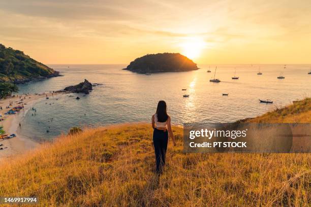 a woman in white t-shirt jeans stands on a mountain with yellow grass overlooking yanui beach, the boat and the mountains below. and there is sunlight - beach morning glory stock pictures, royalty-free photos & images