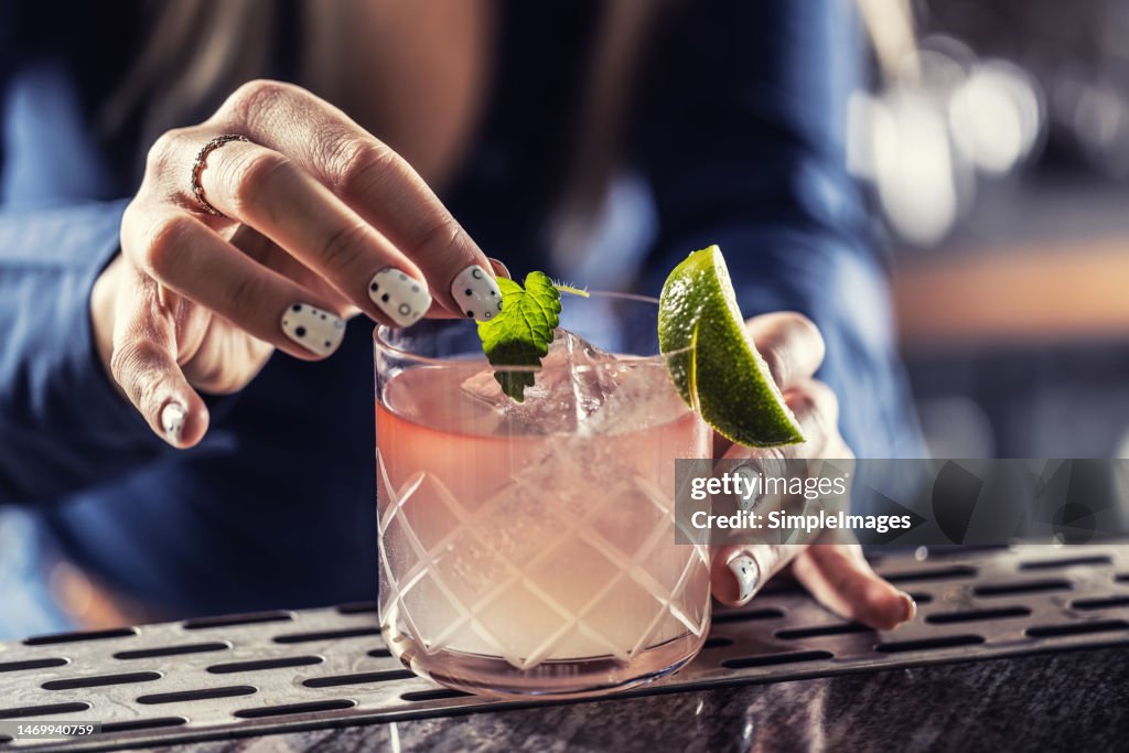 Bartenders hands prepare Margarita cocktail on the bar counter, she decorates the drink with a mint leaf.