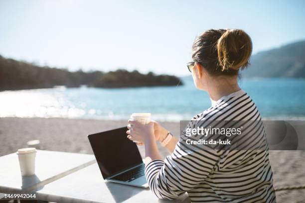 working outdoors. middle aged woman using laptop sitting on the beach and drinking coffee - café da internet stock pictures, royalty-free photos & images