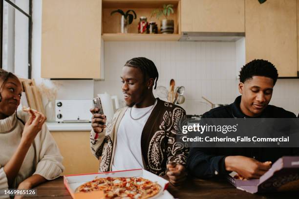 a young man photographs a female with his phone, as she eats a slice of pizza. - dining table icon stock pictures, royalty-free photos & images