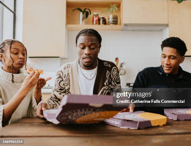 three young people sit in a stylish kitchen and share pizzas directly from the box - chewing with mouth open stock pictures, royalty-free photos & images