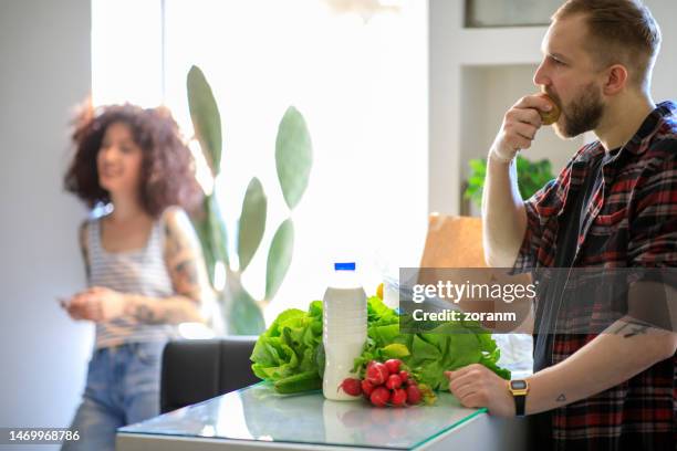 junger mann isst apfel an der theke zu hause, zurück von der morgendlichen besorgung - pflanzliche ernährung stock-fotos und bilder
