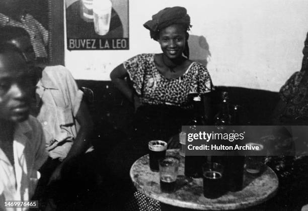 Woman and men sitting in a bar, various drinks and bottles are on their table, Leopoldville, Congo, 1950. There is an advertising poster 'Buvez La...