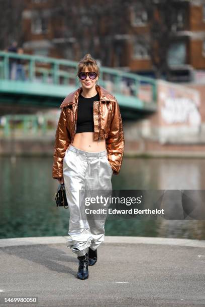 Guest wears black sunglasses, gold large earrings, a black cropped top, a brown shiny metallic shiny leather cropped jacket, silver shiny metallic...