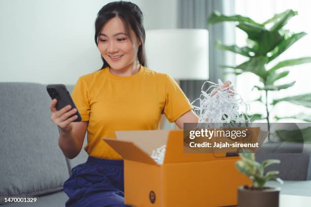 asian woman unpacking carton box happy excited and using smartphone, sitting on sofa in living room at home. - receiving feedback stock pictures, royalty-free photos & images