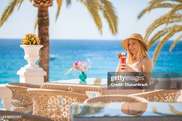 young blonde woman looking at the camera sitting in the beach restaurant on a table drinking delightfully a red cocktail with the turquoise sea in the background - blue white summer hat background stock pictures, royalty-free photos & images