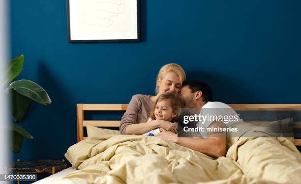 beautiful happy parents cuddling with their daughter in bed - three people in bed stock pictures, royalty-free photos & images