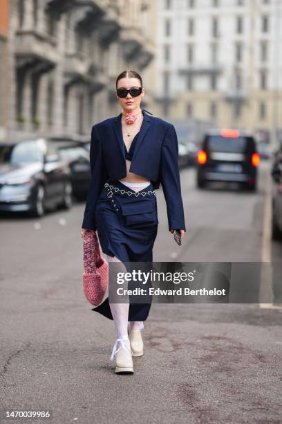 Guest wears black sunglasses, a pink rose flower necklace, a navy blue cropped jacket, a matching navy blue asymmetric long and short skirt, white...
