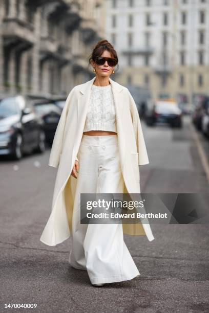 Guest wears black futurist sunglasses, gold earrings, a white braided wool cropped pullover, a white wool long coat, high waist white oversized wide...