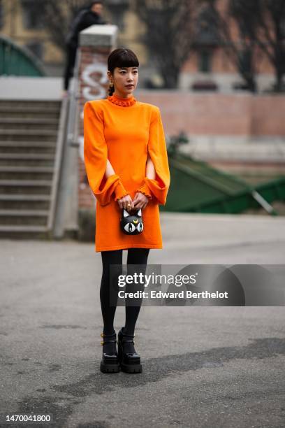 Guest wears gold earrings, a neon orange ruffled high neck / cut-out long sleeves / short dress, a black shiny leather and white details handbag in...