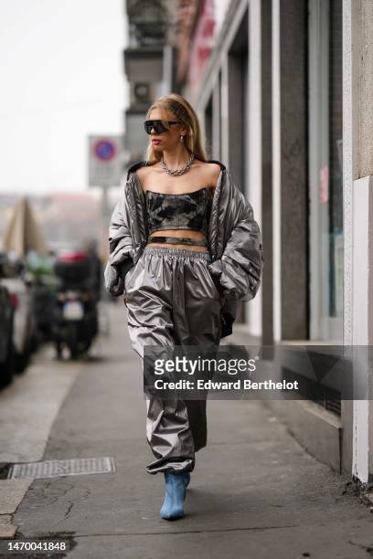 Guest wears black futurist sunglasses, silver earrings, a silver large chain necklace, a silver nylon puffer cropped jacket, matching silver nylon...