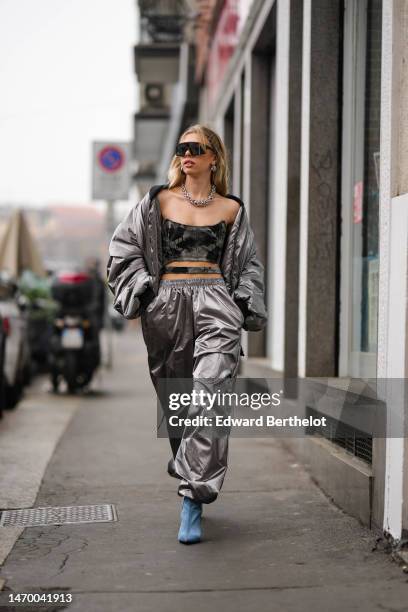 Guest wears black futurist sunglasses, silver earrings, a silver large chain necklace, a silver nylon puffer cropped jacket, matching silver nylon...