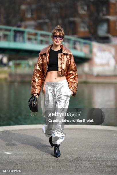 Guest wears black sunglasses, gold large earrings, a black cropped top, a brown shiny metallic shiny leather cropped jacket, silver shiny metallic...