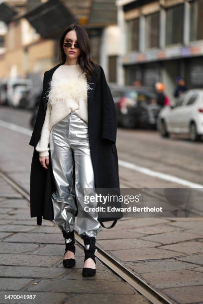 Guest wears black sunglasses, a white wool with embroidered feathers pullover, a black long coat, silver shiny varnished leather large pants, black...