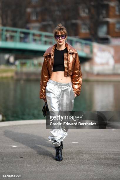 Guest wears black sunglasses, gold large earrings, a black cropped top, a brown shiny metallic shiny leather cropped jacket, silver shiny metallic...