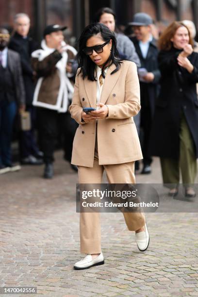 Guest wears black sunglasses, a gold necklace, a white shirt, a beige blazer jacket, beige suit pants, white shiny leather loafers , outside Gucci,...