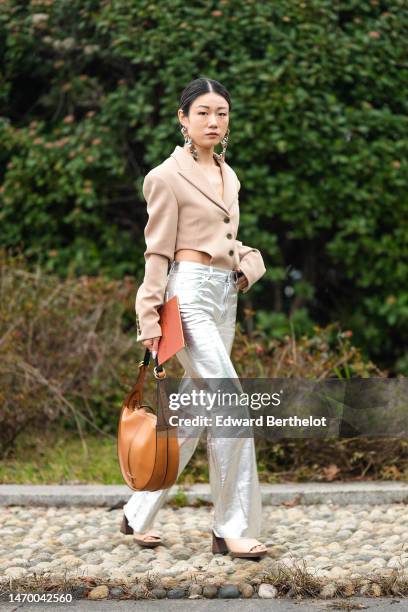 Guest wears silver large pendant earrings, a beige cut-out waist blazer jacket body top, silver shiny leather flared pants, a camel shiny leather...