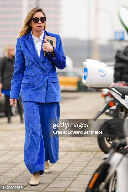 Guest wears black sunglasses, earrings, a white shirt, a royal blue velvet blazer jacket, matching velvet ribbed large suit pants, beige matte...