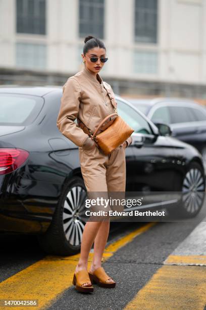 Guest wears black sunglasses, gold earrings, a beige zipper leather bomber coat, a camel shiny leather crossbody bag, beige matching shiny leather...