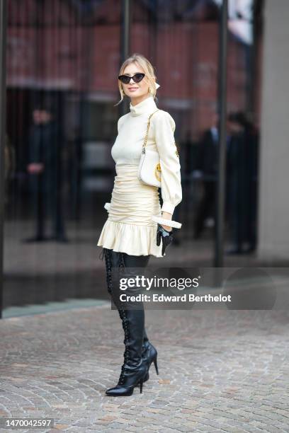 Xenia Adonts wears black vintage sunglasses, a white latte turtleneck long puffy sleeves pullover, a beige satin / silk ruffled short skirt, a white...