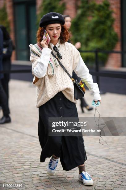 Guest wears a black felt wool with embroidered Paris logo pattern beret, black sunglasses, a white shirt, a white latte braided wool sleeveless...