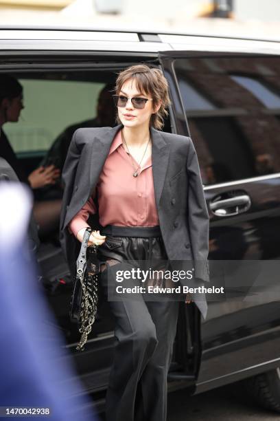 Guest wears black sunglasses, a pale pink silk shirt, a dark gray blazer jacket, a black shiny varnished leather nailed / studded handbag, a gold...