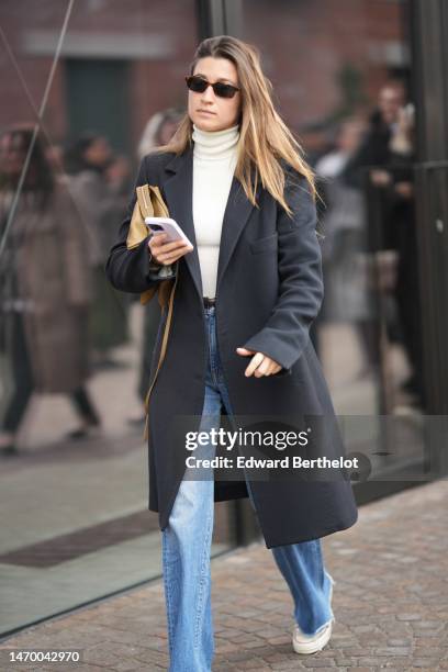 Guest wears black sunglasses, a white turtleneck pullover, a dark gray coat, a beige leather handbag, blue denim faded large pants, white sneakers ,...