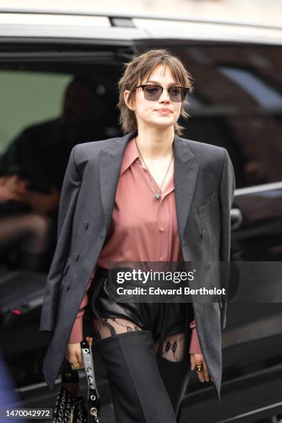 Guest wears black sunglasses, a pale pink silk shirt, a dark gray blazer jacket, a black shiny varnished leather nailed / studded handbag, a gold...