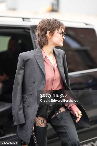 Guest wears black sunglasses, a pale pink silk shirt, a dark gray blazer jacket, a black shiny varnished leather nailed / studded handbag, a gold...